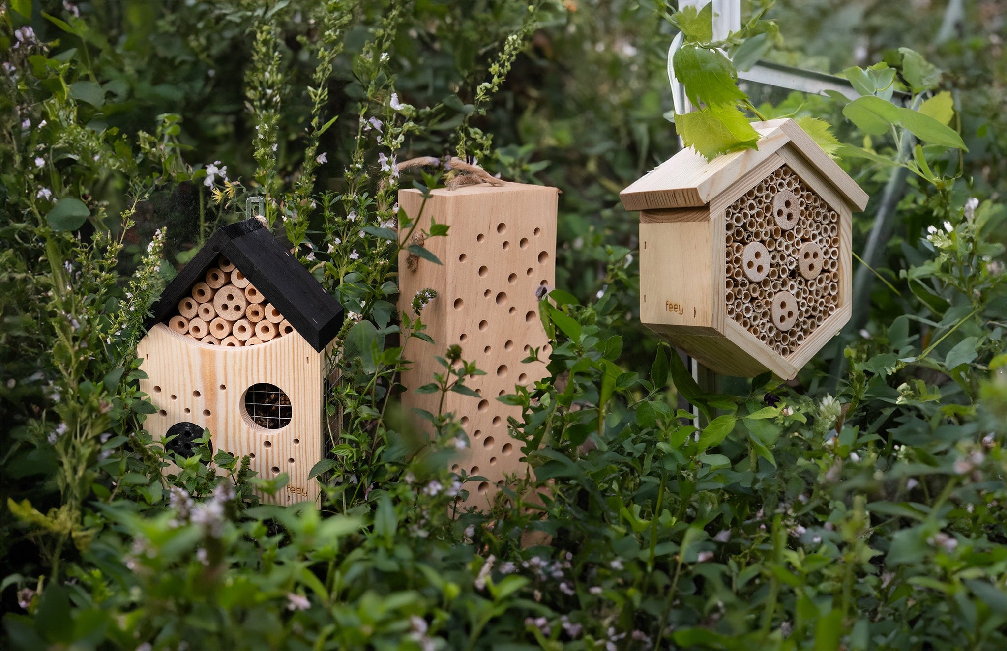 Drei Bienenhotels unterschiedlicher Formen aus Holz und Bambus, eingebettet zwischen üppigen grünen Pflanzen in einer Gartenumgebung.