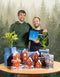 Zwei lächelnde Männer stehen hinter einem Tisch mit getopften Zimmerpflanzen und Miniaturhäusern. Einer hält eine blaue Pflanzen Adventskalender | NIKIN-Special (Dschungel-Version) Box. Im Hintergrund ist eine verschneite, immergrüne Waldszene zu sehen.