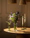 Der Purpurpoesie-Strauß aus violetten und weißen Blumen in einer klaren Glasvase steht auf einem runden Holztisch neben zwei hohen Kerzen und einer kleinen grünen Vase mit weißen Blüten, die alle von warmem Licht beleuchtet werden.