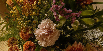 Nahaufnahme eines Blumenarrangements mit rosafarbenen Nelken, gelben knopfartigen Blüten, lila Statice und orangefarbenen Chrysanthemen mit grünem Laub.