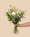 Eine Hand hält den Strauß "Wolkige Grüße" mit weißen Rosen, hellrosa Knospen, lila Blüten, gelben Akzenten und grünen Blättern vor einem schlichten beigen Hintergrund.