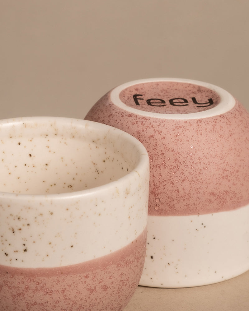 Zwei kleine Tassen aus dem Keramikset „Variado“ mit einem gesprenkelten rosa-weißen Design stehen auf einer neutralen beigen Oberfläche. Eine Tasse ist umgedreht und zeigt auf ihrem Boden den Markennamen „feey“ und erinnert an entzückende Keramiktöpfe, die für die Präsentation von Jungpflanzen gefertigt wurden.