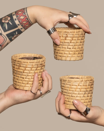 Drei tätowierte, ringgeschmückte Hände halten die Rattan Babypflanzen-Körbe 'Calen' (3 Stk.) vor einem schlichten beigen Hintergrund.
