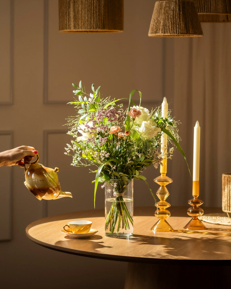 Eine Hand gießt Tee aus einer Teekanne in eine Tasse auf einem runden Holztisch, der mit Wolkige Grüsse, einem hohen Blumenstrauß und zwei brennenden gelben Kerzen in eleganten Haltern geschmückt ist. Das warme Licht zieht die Aufmerksamkeit auf sich und schafft eine gemütliche Atmosphäre.