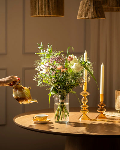 Eine Hand gießt Tee aus einer Teekanne in eine Tasse auf einem runden Holztisch, der mit Wolkige Grüsse, einem hohen Blumenstrauß und zwei brennenden gelben Kerzen in eleganten Haltern geschmückt ist. Das warme Licht zieht die Aufmerksamkeit auf sich und schafft eine gemütliche Atmosphäre.