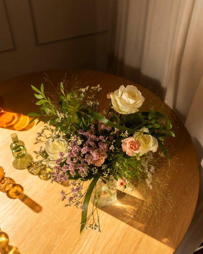 Der Blumenstrauß "Wolkige Grüsse" mit weißen und zartrosa Rosen, violetten Blumen und Grünzeug steht auf einem runden Holztisch in sanfter Beleuchtung. Glasflaschen in der Nähe lenken die Aufmerksamkeit auf das Arrangement.