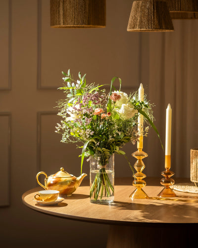 Ein runder Holztisch mit Wolkige Grüsse, zwei hohe gelbe Kerzen in Glashaltern, eine goldene Teekanne und eine passende Teetasse, alles beleuchtet von warmem Sonnenlicht, das jedes Detail hervorhebt.