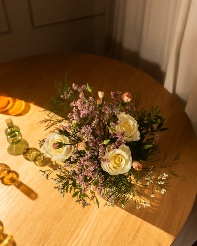 Der Wolkige Grüsse Strauss mit weissen Rosen, rosa Blumen und Grünzeug steht auf einem runden Holztisch im warmen Sonnenlicht, umgeben von dekorativen Glasakzenten und luftigen Vorhängen im Hintergrund.
