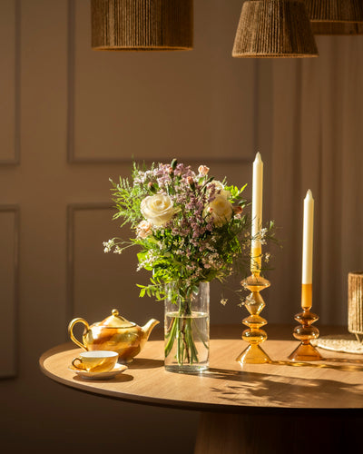 Auf einem sonnenbeschienenen Holztisch stehen "Wolkige Grüße", ein Strauß weißer und rosafarbener Blumen, neben bernsteinfarbenen Kerzenhaltern und einer goldenen Teekanne. Geflochtene Lampenschirme werfen ein weiches, pastellfarbenes Licht, das diese friedliche Szene unterstreicht.