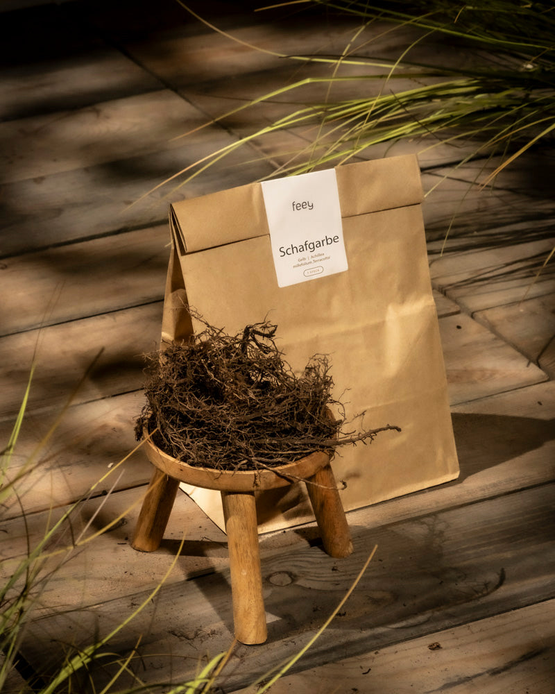 Eine braune Papiertüte mit der Aufschrift „feey Schafgarbe (orange-braun)“ liegt auf Holzdielen neben einem Hocker mit getrockneten Achillea millefolium-Kräutern, in der Nähe befindet sich grünes Gras.