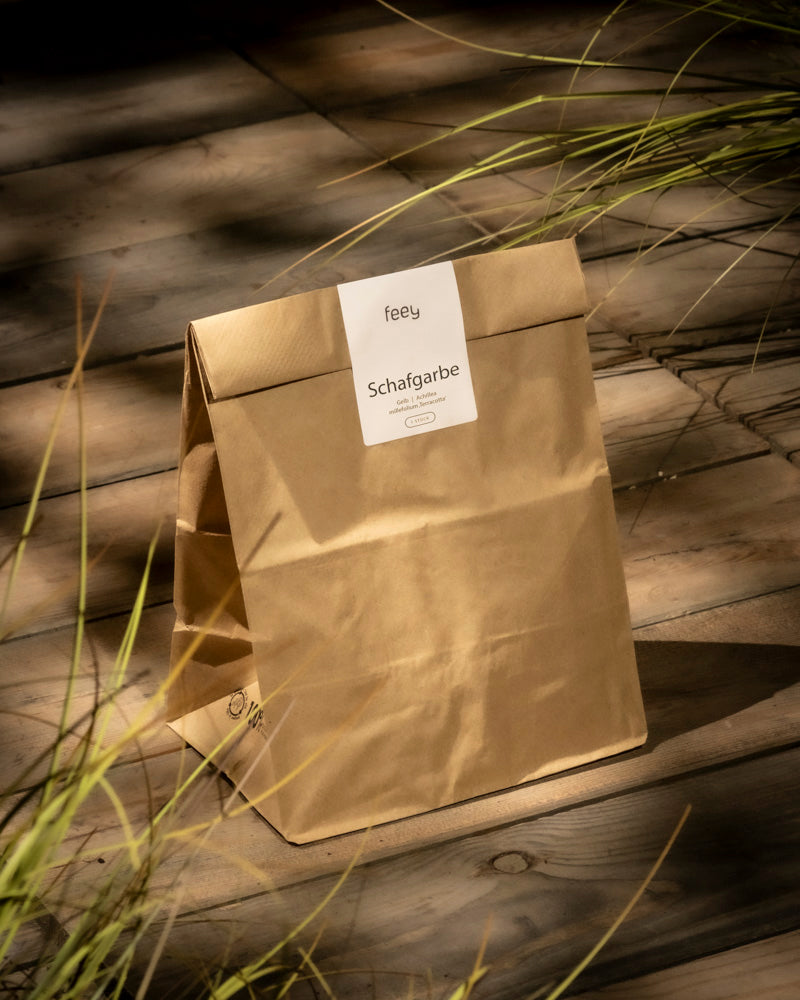 Eine braune Papiertüte mit der Aufschrift „feey Schafgarbe (orange-braun)“ steht aufrecht auf einer sonnenbeschienenen Holzoberfläche, im Vordergrund ist grünes Gras zu sehen – was die natürliche Herkunft von Achillea millefolium unterstreicht.