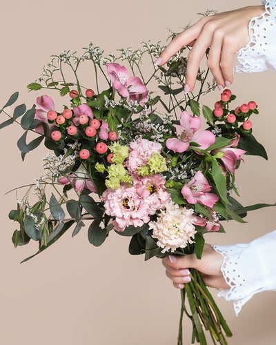 Eine Hand hält den Ballerina-Strauß - rosa und weiße Blumen, grüne Blätter und rote Beeren - vor einem beigen Hintergrund, während eine andere Hand mit Spitzenmanschetten die Blüten sanft von oben arrangiert.