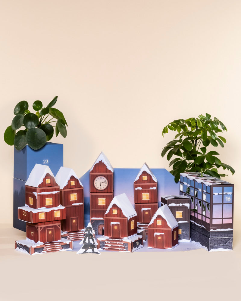 Ein Pflanzen-Adventskalender in Form eines verschneiten Dorfes mit leuchtenden Fenstern steht auf einem Tisch, flankiert von zwei pflegeleichten Topfpflanzen im Hintergrund.