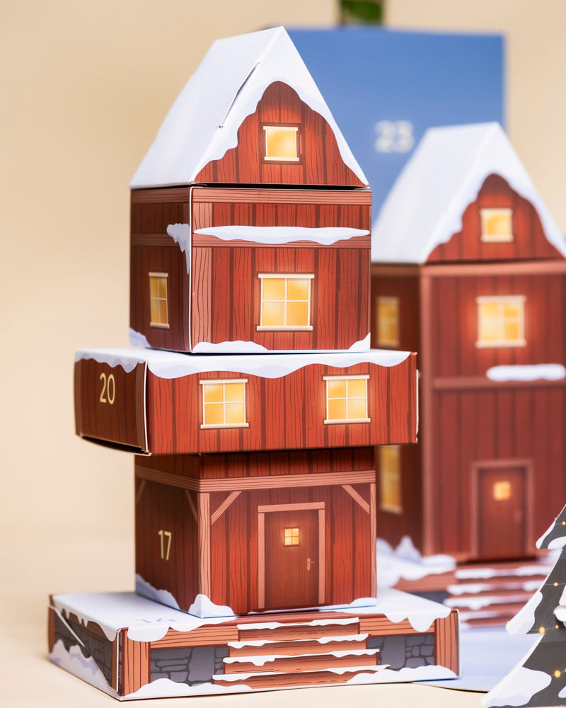 Ein festlicher Pflanzen-Adventskalender in Form eines schneebedeckten Holzhauses mit beleuchteten Fenstern und nummerierten Türen 17, 20 und 23. Ein weiteres ähnliches Haus ist im Hintergrund zu sehen.