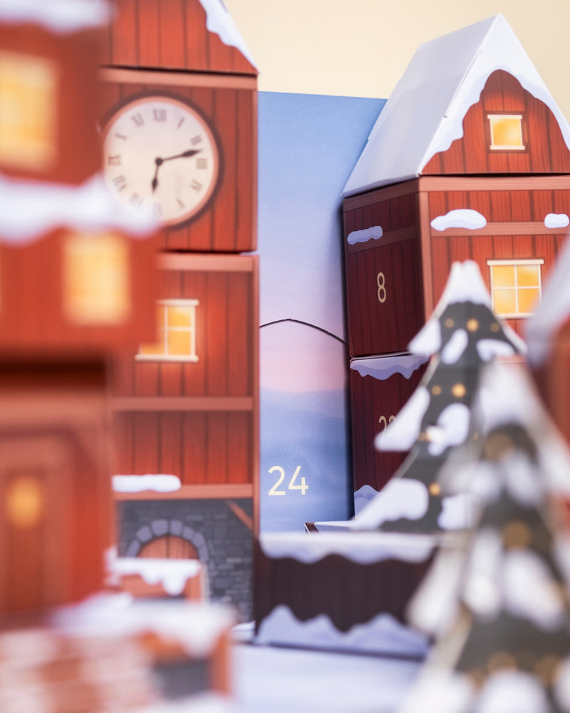 Eine festliche Szene mit roten Holzhäusern, verschneiten Dächern, einem Uhrenturm und einem Weihnachtsbaum weist auf die Zahl 24 und den Pflanzen-Adventskalender hin, der hinter jedem Türchen echte Pflanzen zeigt.