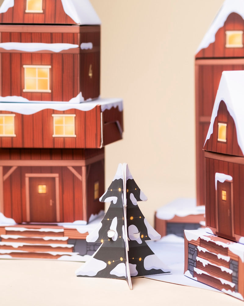 Ein festlicher Weihnachtsbaum aus Pappe mit gelben Lichtern und weißem Schnee steht vor roten, verschneiten Häusern mit leuchtenden Fenstern - ein bezaubernder Dekorationsakzent, der in dieser Saison mit dem Pflanzen Adventskalender kombiniert werden kann.