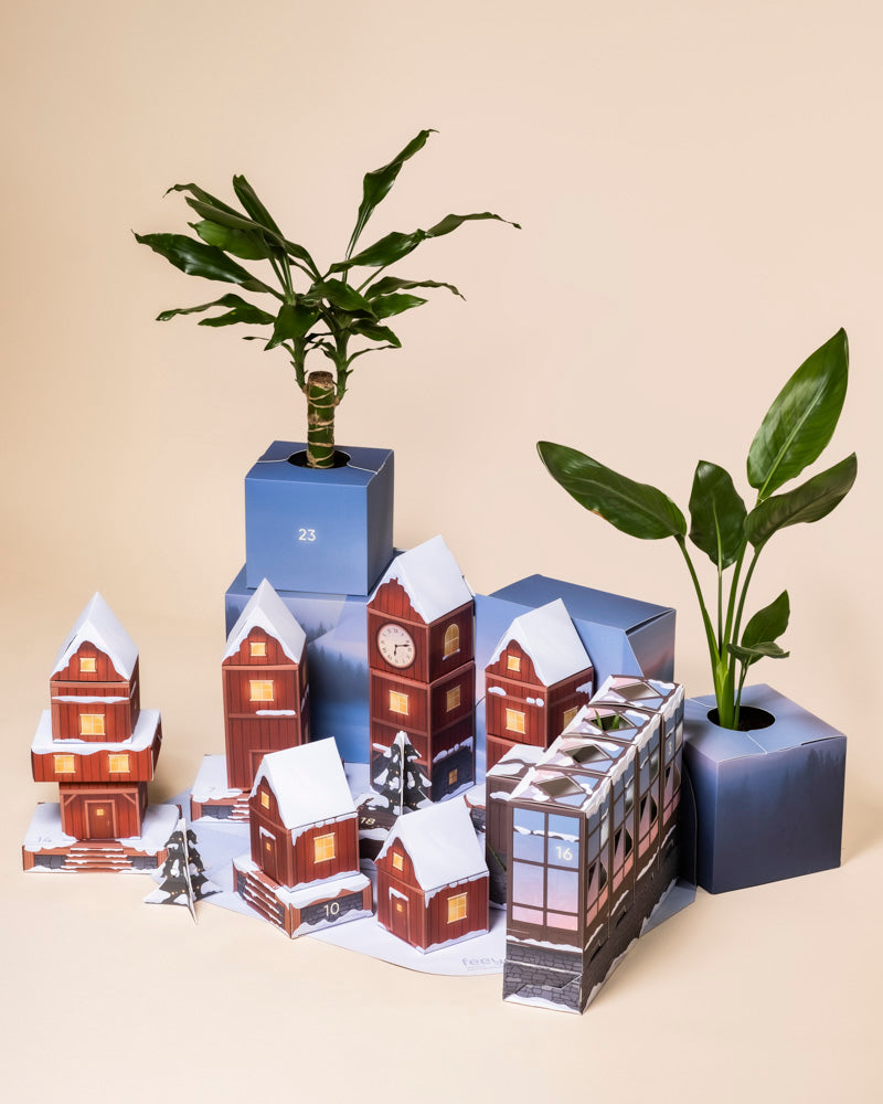 Eine festliche Szene mit kleinen verschneiten Modellhäusern und Tannenbäumen, eingebettet in blaue geometrische Pflanzgefäße mit echten und pflegeleichten Pflanzen, hebt den Pflanzen-Adventskalender vor einem neutralen Hintergrund hervor.