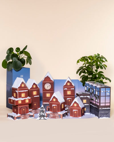 Ein Pflanzen-Adventskalender in Form eines verschneiten Dorfes mit leuchtenden Fenstern steht auf einem Tisch, flankiert von zwei pflegeleichten Topfpflanzen im Hintergrund.