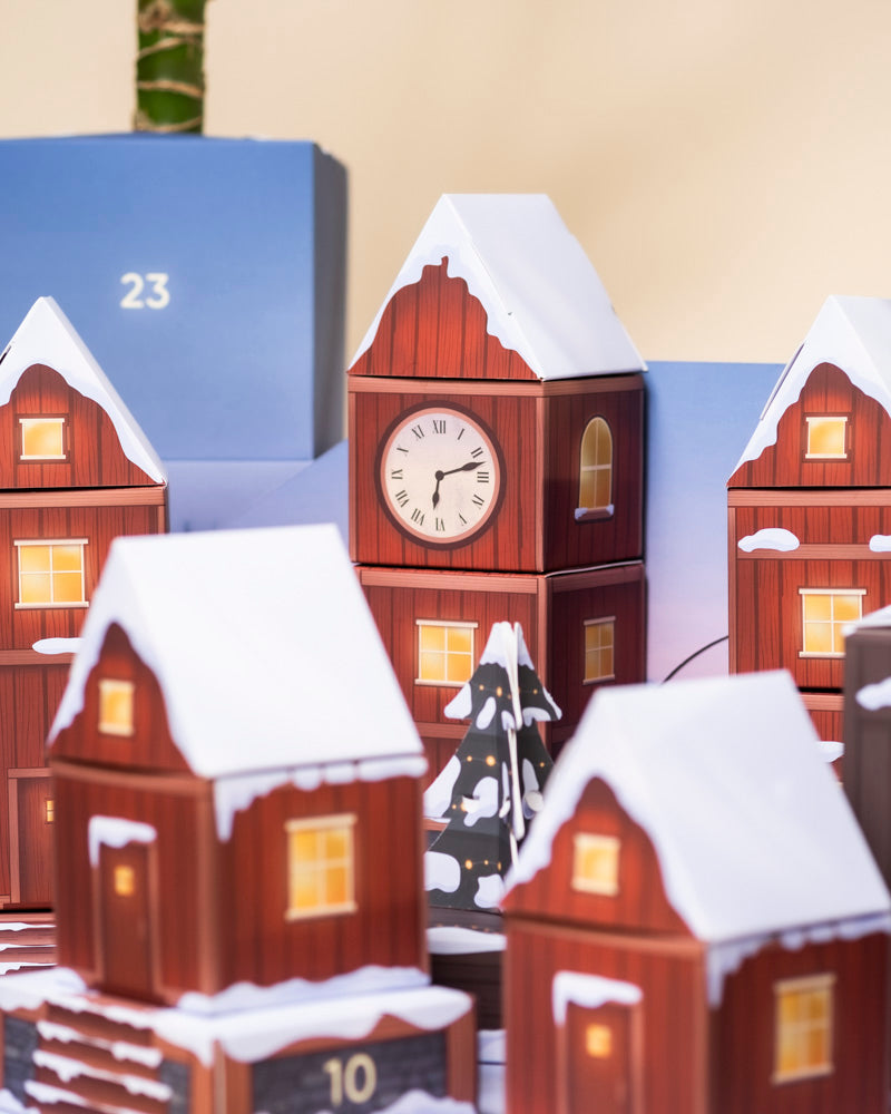 Eine Nahaufnahme einer festlichen Szene mit dekorativen Häusern und einem verschneiten Uhrenturm vor der Zahl 23, die den Charme des Pflanzen-Adventskalenders einfängt.