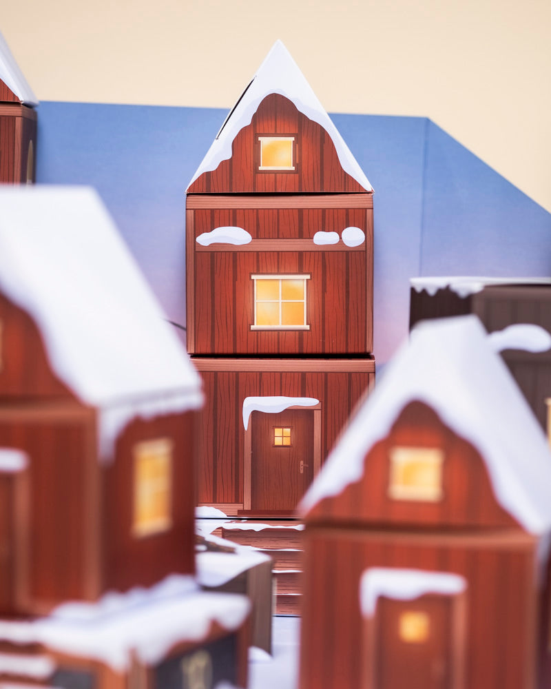 Ein gemütliches Holzhaus mit schneebedecktem Dach und leuchtenden Fenstern sorgt für eine warme Atmosphäre. Im Fenster bringt ein Pflanzen-Adventskalender echte Pflanzen und einen Hauch von Natur in die winterliche, zartblaue Kulisse.