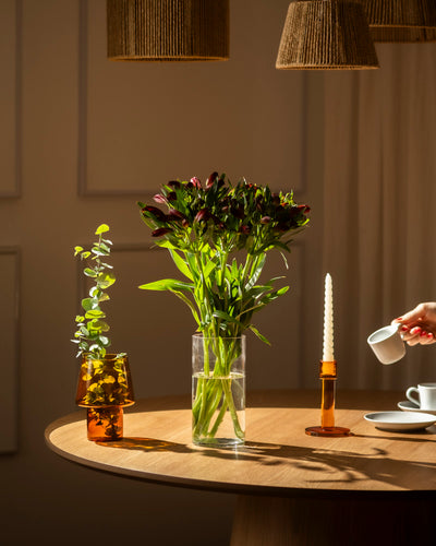 Auf einem runden Holztisch stehen eine Lieblingsvase mit 10 roten Alstroemeria, ein braunes Glasgefäß mit Grünzeug, ein orangefarbener Kerzenhalter mit einer weißen Kerze und eine Hand, die bei warmer Beleuchtung Milch in eine Tasse gießt.