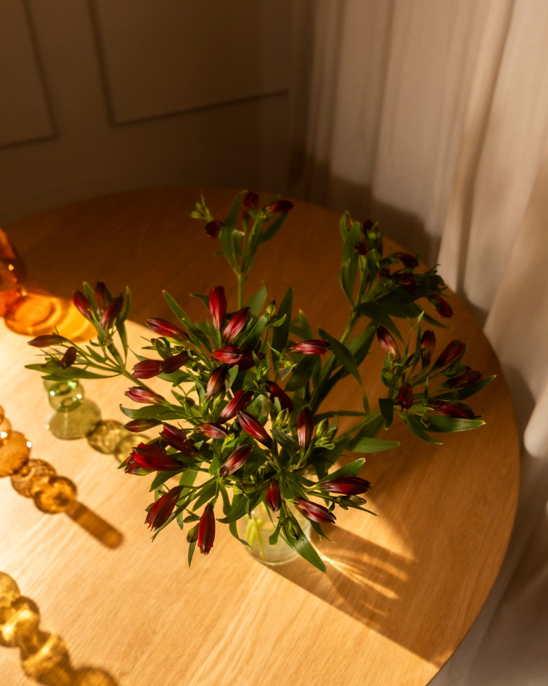 Ein Strauß Alstroemeria rot (10 Stk.) in Ihrer Lieblingsvase steht auf einem runden Holztisch, der durch weiße Vorhänge in warmes Sonnenlicht getaucht ist, mit dekorativen Glasornamenten in der Nähe.