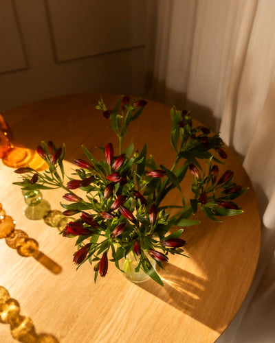 Ein Strauß Alstroemeria rot (10 Stk.) in Ihrer Lieblingsvase steht auf einem runden Holztisch, der durch weiße Vorhänge in warmes Sonnenlicht getaucht ist, mit dekorativen Glasornamenten in der Nähe.
