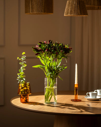 Eine Lieblingsvase aus klarem Glas enthält einen Strauß Alstroemeria rot (10 Stk.) mit grünen Blättern, der auf einem Holztisch neben einer gedrehten weißen Kerze in einem bernsteinfarbenen Halter, einer kleinen bernsteinfarbenen Vase mit Grünzeug und zwei weißen Teetassen steht.