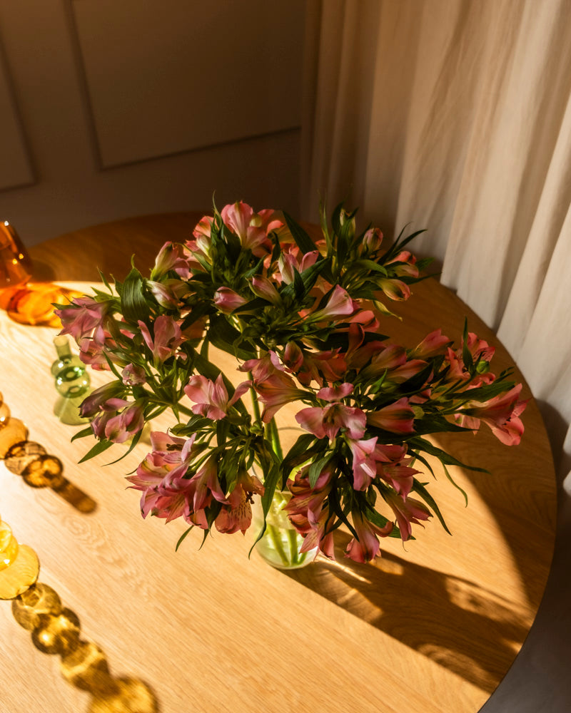 Ein Strauß Alstroemeria rosa (10 Stk.) steht in einer Vase auf einem Holztisch. Das Sonnenlicht fällt ein und wirft weiche Schatten. An der Seite des Tisches sind dekorative Glasobjekte angeordnet.