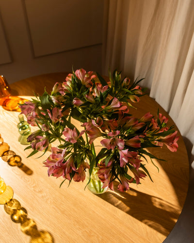 Ein Strauß Alstroemeria rosa (10 Stk.) steht in einer Vase auf einem Holztisch. Das Sonnenlicht fällt ein und wirft weiche Schatten. An der Seite des Tisches sind dekorative Glasobjekte angeordnet.