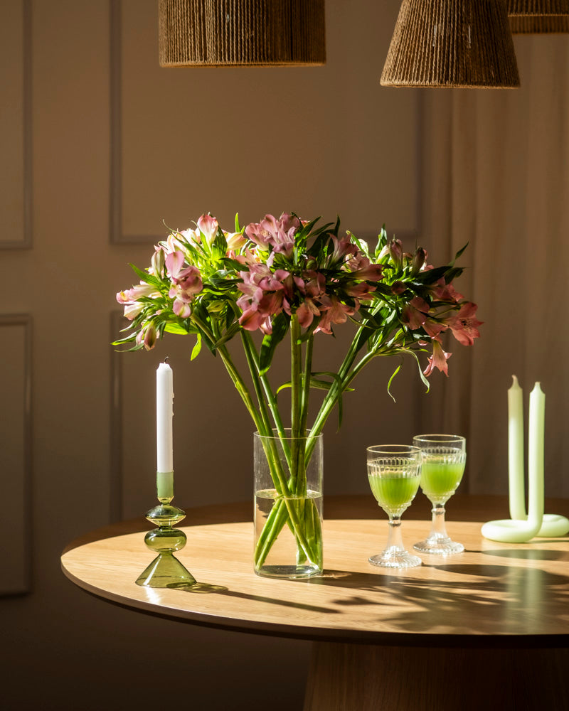 Eine klare Glasvase mit Alstroemeria rosa (10 Stk.) steht auf einem runden Holztisch, daneben eine brennende weiße Kerze in einem grünen Halter, zwei grüne Getränke in eleganten Gläsern und zwei nicht brennende grüne Kerzen. Warmes Licht taucht die Szene.