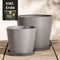 Zwei graue Pflanzkübel-Set Amsterdam (40, 30) unterschiedlicher Größe stehen nebeneinander auf einer Holzfläche. Jeder Topf verfügt über einen passenden Untersetzer und ein effizientes Drainage-System. In der oberen linken Ecke ist ein kleines Bild eines Erdsacks mit dem Text „Inkl. Erde“ zu sehen, der darauf hinweist, dass Erde im Lieferumfang enthalten ist.