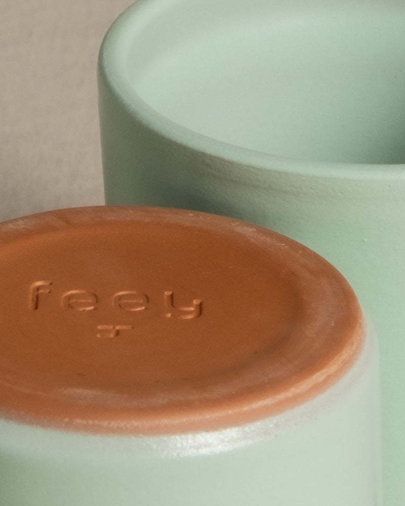 Nahaufnahme von zwei Keramiktassen. Eine Tasse ist verkehrt herum ausgestellt und zeigt einen terrakottafarbenen Boden mit dem eingeprägten Wort „feey“. Die übrigen Tassen, die perfekt für Keramik Babypflanzen-Topf „Sencillo“ oder Feey-Babypflanzen geeignet sind, haben einen hellgrünen Farbton.