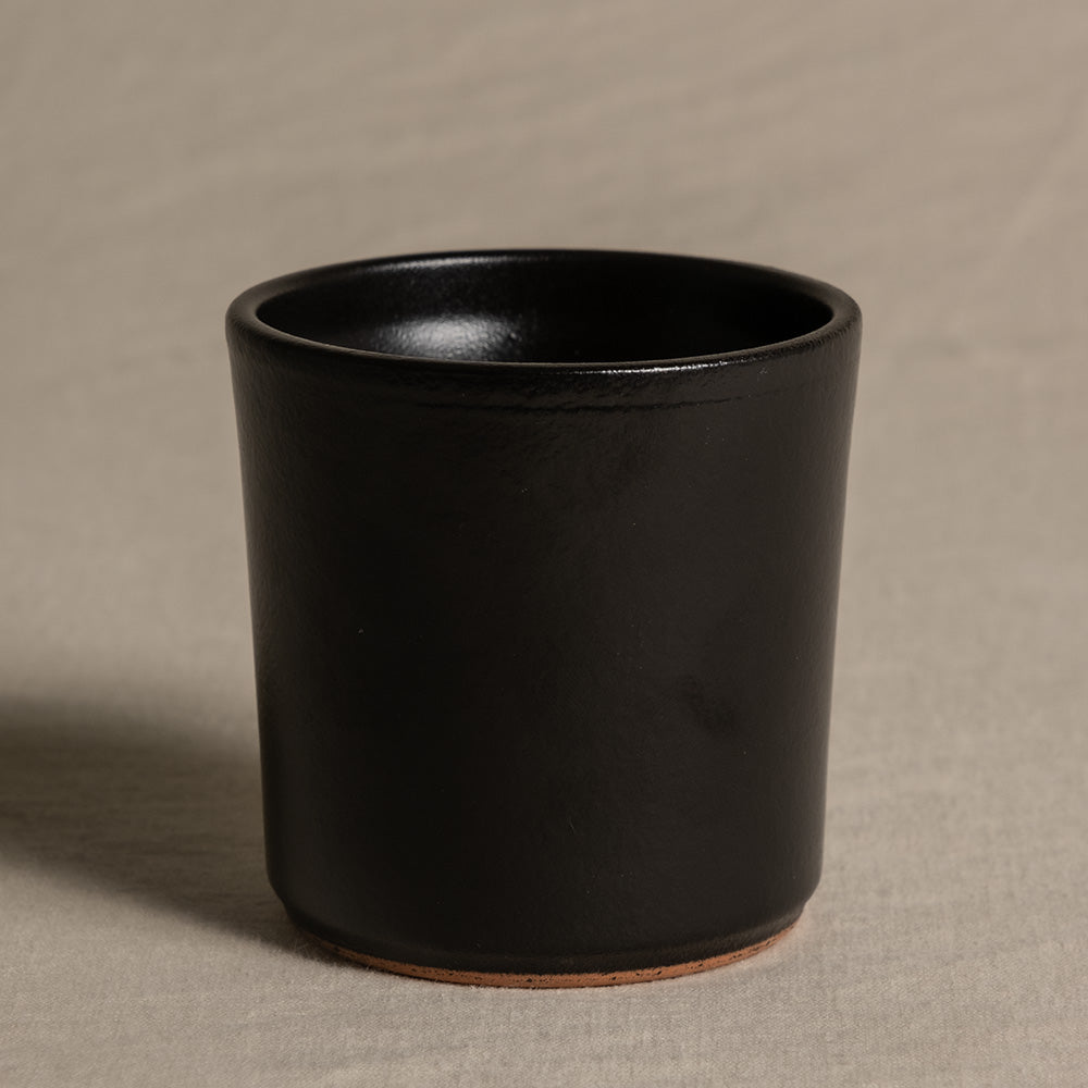 Ein schwarzer Keramik-Babypflanzen-Topf „Sencillo“ mit minimalistischem Design steht auf einer hellgrauen strukturierten Oberfläche. Der Topf ist zylindrisch und hat eine glatte, matte Oberfläche. Die Innenseite sieht genauso aus wie die Außenseite und am Boden ist ein kleiner Teil natürlichen Tons sichtbar.