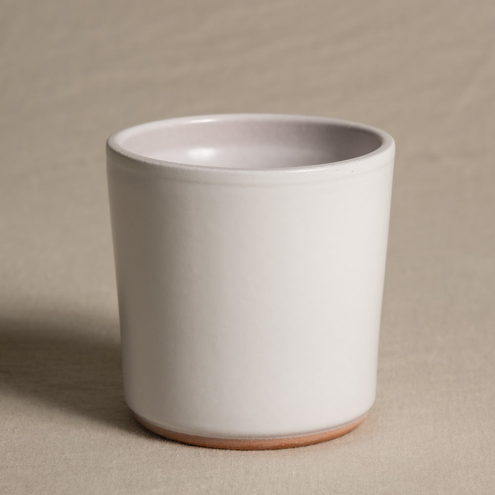 Eine minimalistische weiße Keramiktasse mit glatter, matter Oberfläche und leicht konischer Form. Sie ähnelt einem Keramik-Babypflanzen-Topf „Sencillo“ und steht auf einer flachen Oberfläche, die mit hellem Stoff bedeckt ist. Die Innenseite der Tasse weist einen glänzenden Lavendelton auf.