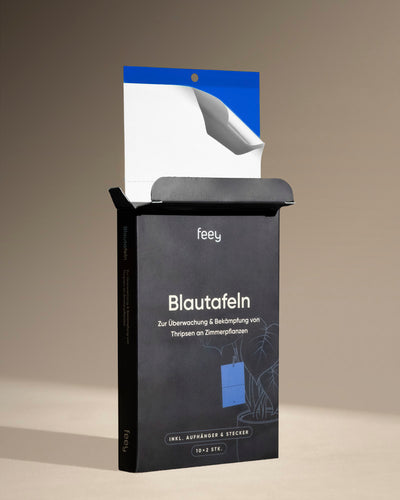 Eine offene schwarze Box mit der Aufschrift „Bekämpfungsset gegen Thripse“ zeigt eine teilweise entfernte blaue Klebefalle mit weißer Rückseite, die zur Überwachung und Bekämpfung von Thripse an Zimmerpflanzen entwickelt wurde und auf einer neutralen Oberfläche steht.