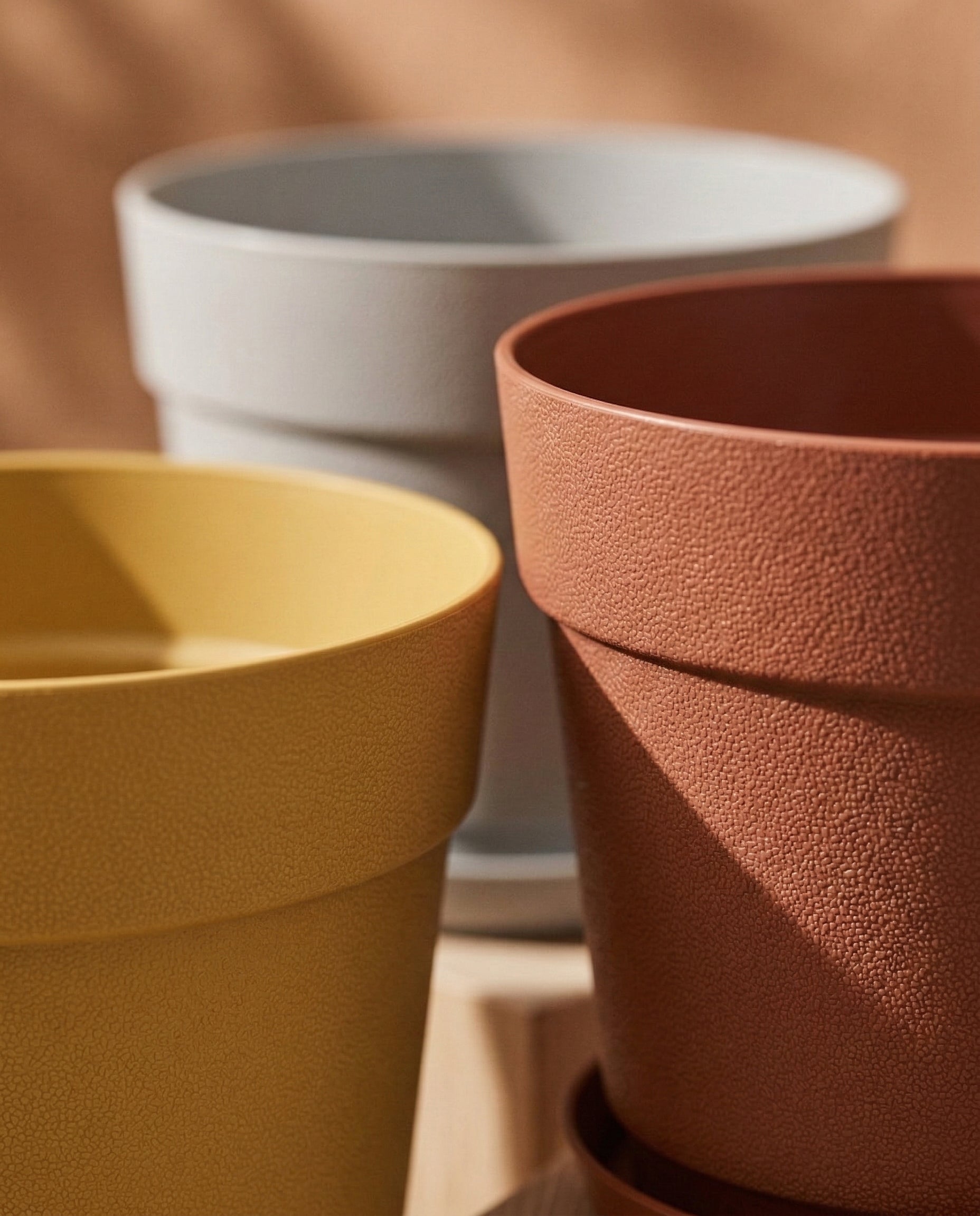 Das Pflanzkübel-Set 'Bloom' (3× 29) besteht aus drei strukturierten Blumentöpfen in Gelb, Terrakotta und Hellgrau aus recyceltem Kunststoff. Diese UV-beständigen Töpfe heben sich von einem sanft verschwommenen bräunlichen Hintergrund ab.