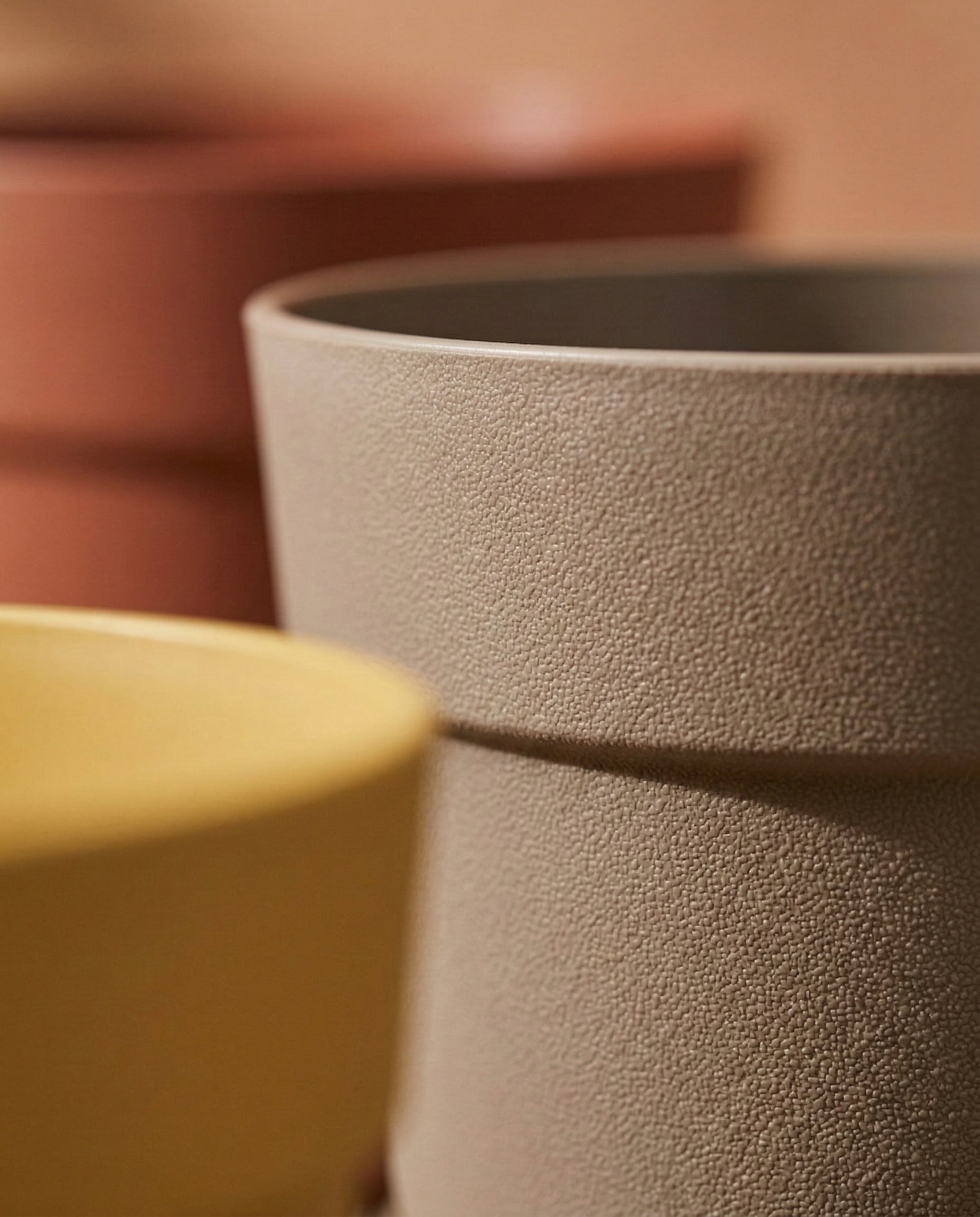 Nahaufnahme des Pflanzkübel-Sets 'Bloom' (3× 29) in Beige, Gelb und Terrakotta, hergestellt aus recyceltem Kunststoff. Diese UV-beständigen Töpfe mit strukturierter Oberfläche sind nebeneinander vor einem weichen, verschwommenen Hintergrund angeordnet.