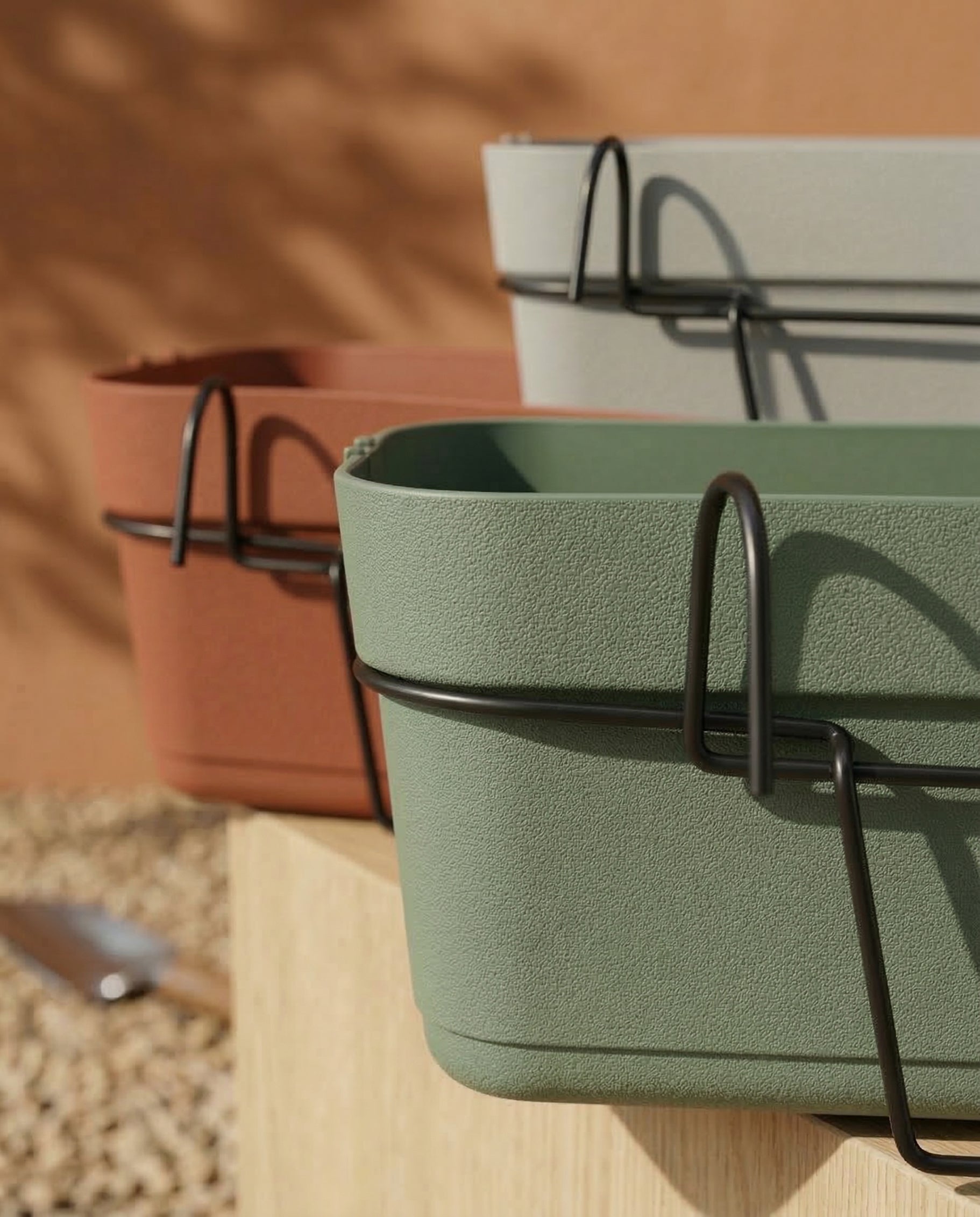 Drei rechteckige Pflanzkästen für Geländer (Blossom | 60 cm) in Grün, Grau und Terrakotta mit schwarzen Metallhaltern und Wasserreservoirs stehen auf einem Holzbalkon, der in Sonnenlicht getaucht ist und eine warme, natürliche Atmosphäre im Freien schafft.