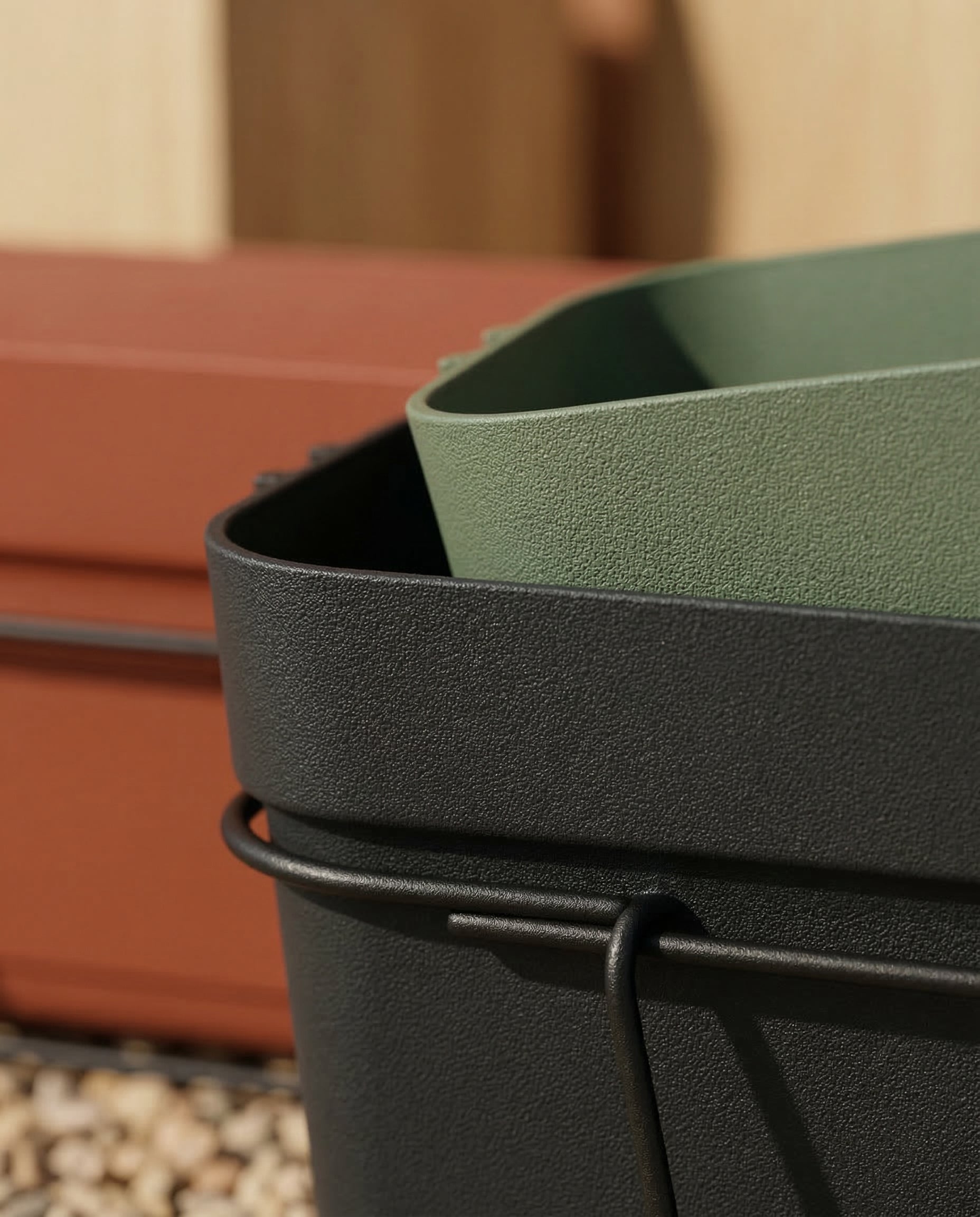 Nahaufnahme von drei gestapelten Pflanzkästen für Geländer (Blossom | 60 cm) in Schwarz, Grün und Terrakotta, ideal für den Balkon, mit Kieselsteinen und Holzoberflächen, die im Hintergrund sanft verschwimmen.