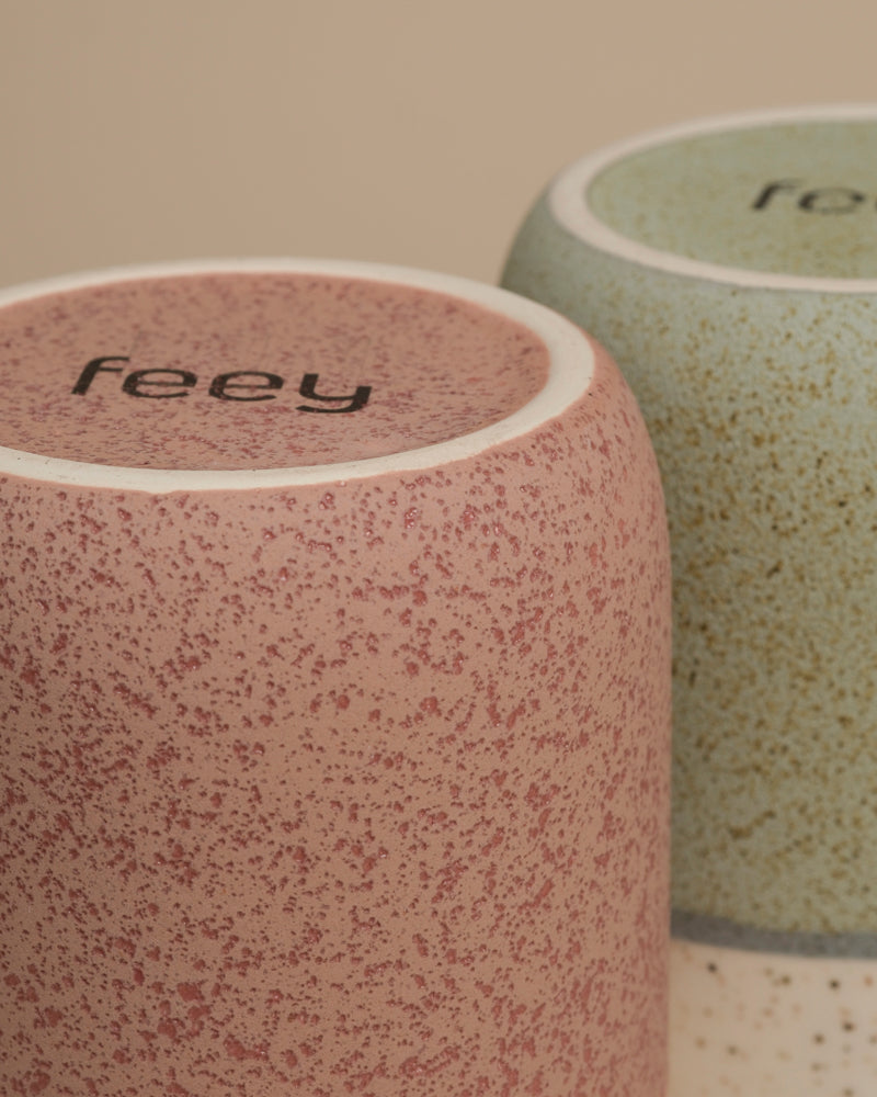 Nahaufnahme von zwei umgedrehten Keramikbechern mit gesprenkelter Textur, einer davon rosa mit "feey" auf dem Boden. Perfekt neben einer feey Keramik-Vase (Variado | 21 cm) oder zwischen Zimmerpflanzen.