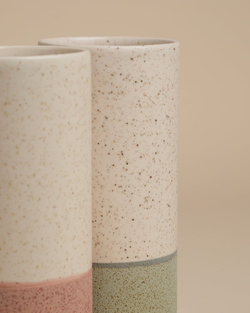 Zwei hohe Keramik-Vasen (Variado | 21 cm), eine mit einem grünlichen und eine mit einem rosafarbenen Ton, haben ein gesprenkeltes cremefarbenes Oberteil. Stilvoll vor einem beigen Hintergrund - ideal als Blumenvase für Zimmerpflanzen oder als eigenständige Dekorationsstücke.