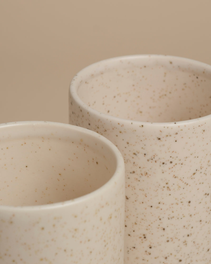 Zwei Keramikbecher mit einer gesprenkelten, cremefarbenen Glasur, die nebeneinander vor einem neutralen beigen Hintergrund stehen, erinnern an die schlichte Eleganz der feey Keramik-Vase (Variado | 21 cm).