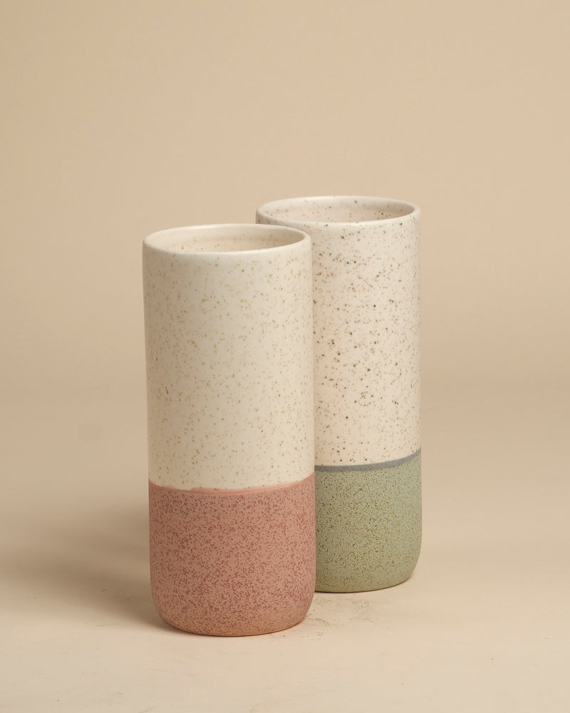 Zwei fey Keramik-Vasen (Variado | 21 cm) mit mattweißer, dezent gesprenkelter Oberfläche - eine hat einen gedeckten rosa Grund, die andere einen grünen. Ideal als Blumenvase für Zimmerpflanzen. Beide sind auf einem beigen Hintergrund abgebildet.