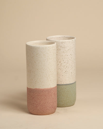 Zwei fey Keramik-Vasen (Variado | 21 cm) mit mattweißer, dezent gesprenkelter Oberfläche - eine hat einen gedeckten rosa Grund, die andere einen grünen. Ideal als Blumenvase für Zimmerpflanzen. Beide sind auf einem beigen Hintergrund abgebildet.