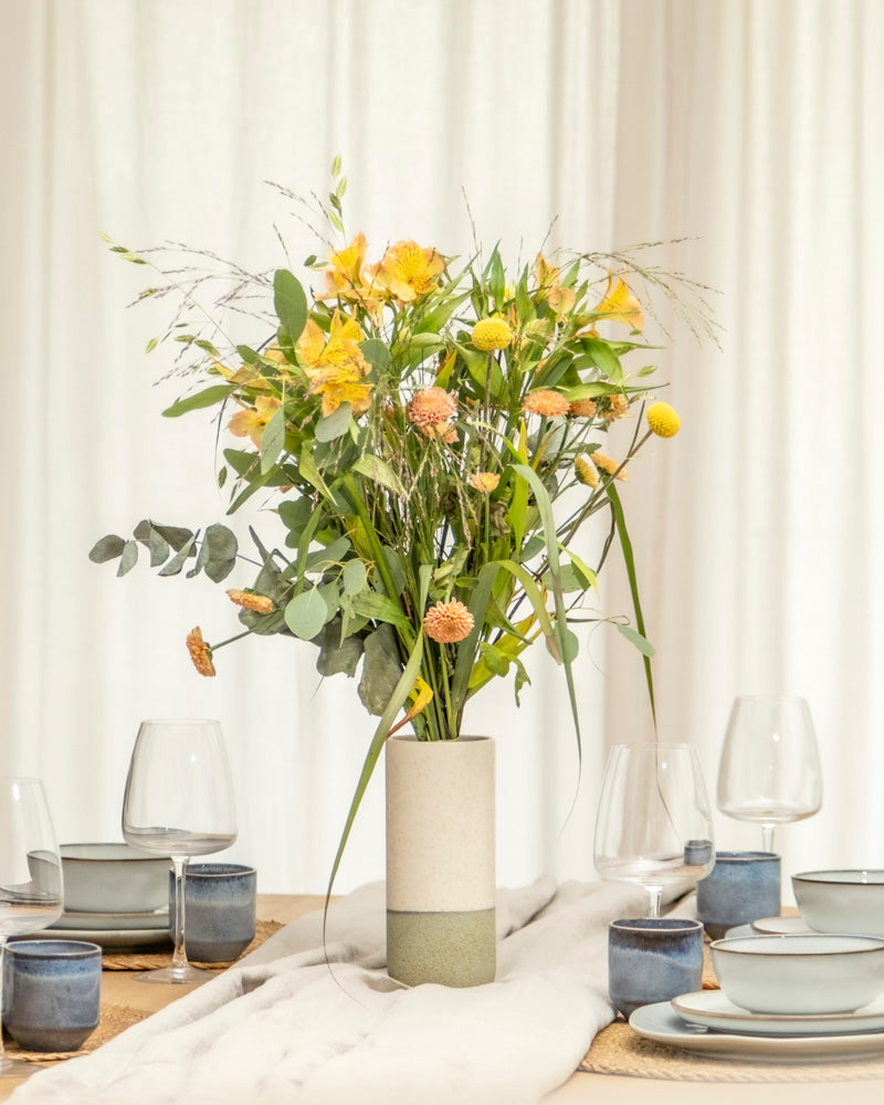 Eine mit gelben und orangefarbenen Blumen gefüllte Keramik-Vase (Variado | 21 cm) steht auf einem mit blauem und beigem Geschirr, Weingläsern und neutraler Bettwäsche gedeckten Esstisch, der von weißen Vorhängen eingerahmt wird.