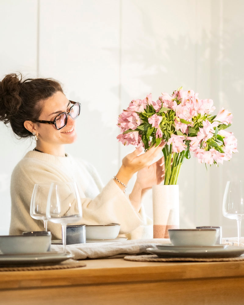 Eine lächelnde Frau mit Brille arrangiert rosa Blumen in einer Keramik-Vase (Variado | 21 cm) auf einem gedeckten Esstisch mit Tellern, Schalen und Weingläsern.