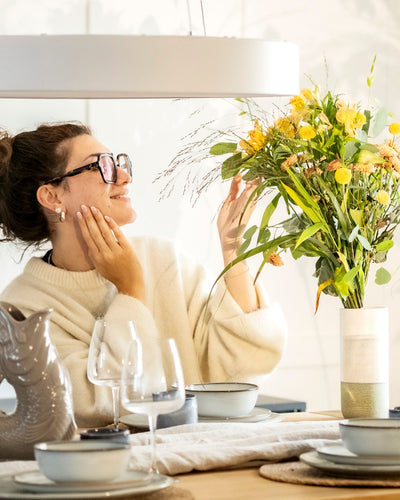 Eine Frau mit Brille und einem gemütlichen weißen Pullover lächelt, während sie gelbe Blumen bewundert, die in der feey Keramik-Vase (Variado | 21 cm) auf einem gedeckten Esstisch mit Gläsern, Tellern und einem fischförmigen Zierkrug arrangiert sind.