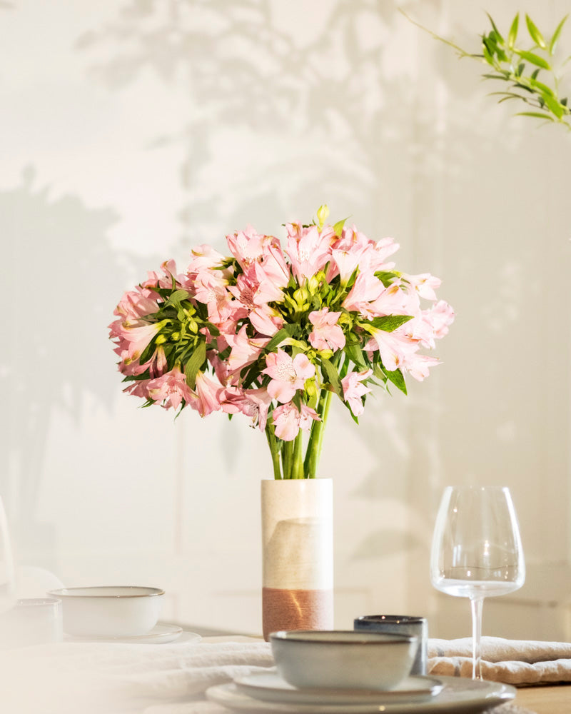 Eine mit hellrosa Blumen gefüllte Keramik-Vase (Variado | 21 cm) steht auf einem Tisch mit Schalen, Tellern und einem Weinglas, während weiches Licht Schatten auf die Wand dahinter wirft.