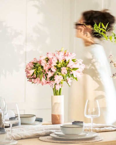 Auf einem gedeckten Tisch steht eine fey Keramik-Vase (Variado | 21 cm) voller pinker Blumen; im Hintergrund geht eine Person mit Brille vorbei.